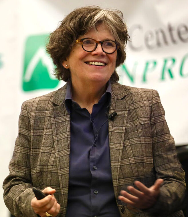 Joan Garry, a woman with short, wavy graying hair and glasses, smiling and gesturing with her hands while speaking at a podium, wearing a plaid blazer and dark shirt against a blurred background with event logos.
